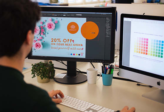 Poste de travail équipé de deux écrans, un collaborateur travaille sur la conception d'une affiche et vérifie les couleurs sur le deuxième écran.